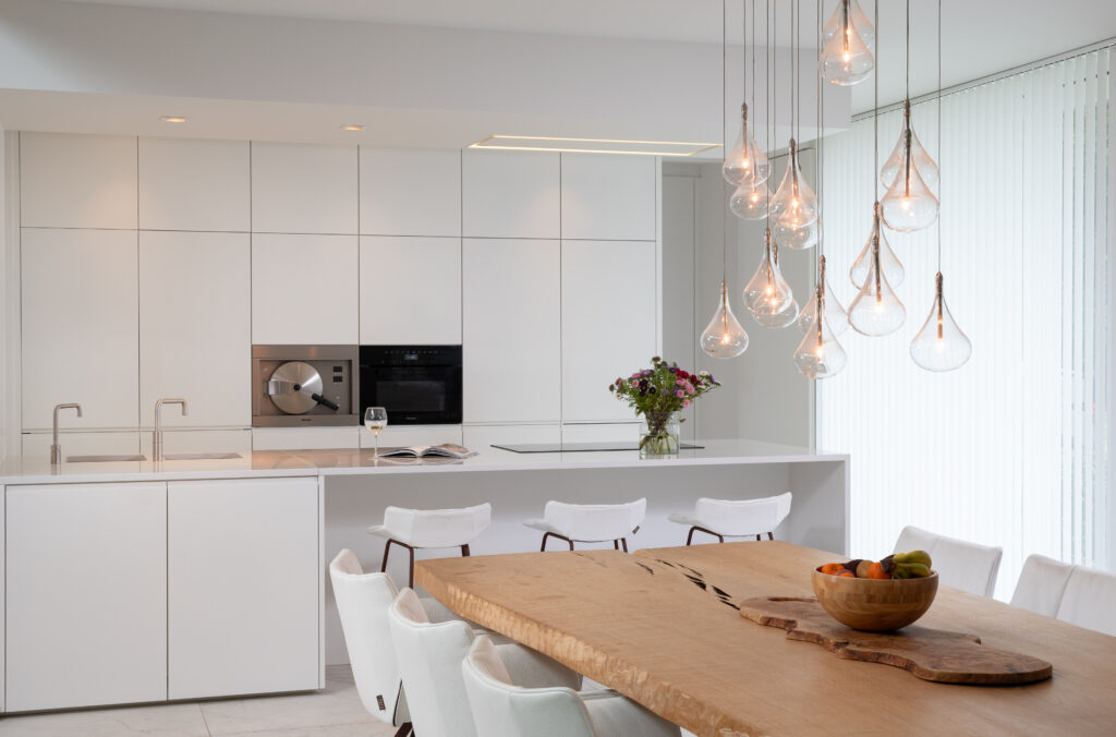 Moderne witte keuken met naadloze kasten, een ruim werkblad met dubbele spoelbakken, en een set glazen hanglampen boven de eettafel. Ingebouwde oven en stoomoven zijn discreet geplaatst.